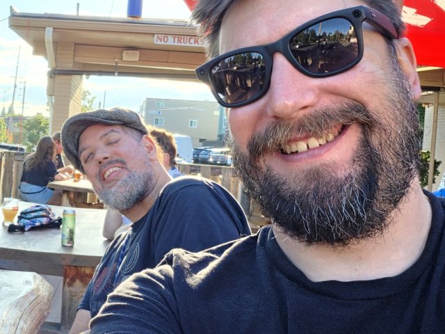 Andrew Dall grins close to the camera in sunglasses while Jordan Remington Williams, in a flat cap, laughs beside him at an outdoor table in Portland.