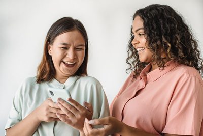 Two women laugh together as they look at a phone one of them is holding.