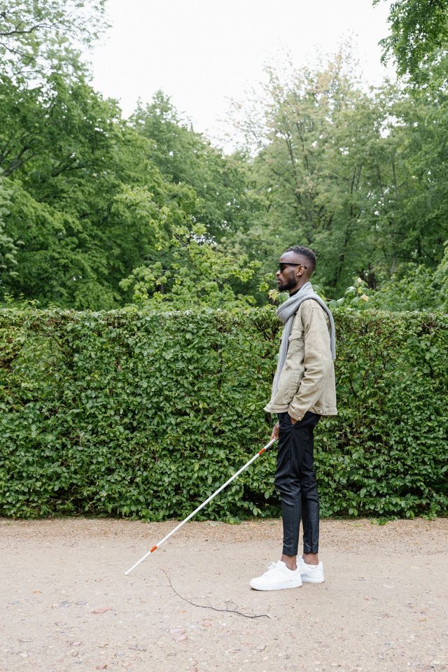 A man stands on a park path in the rain, his white cane extended ahead of him.
