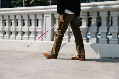 A person mid-stride on a sunlit walkway, a white cane sweeping the ground ahead of them.