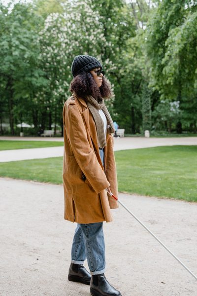 A woman in a long coat walks a park path with a white cane held out ahead of her.