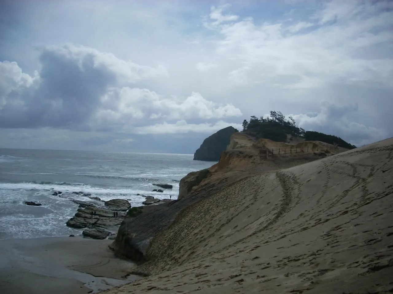 A broad sand dune sloping down to a rocky headland, with surf breaking on the rocks below.